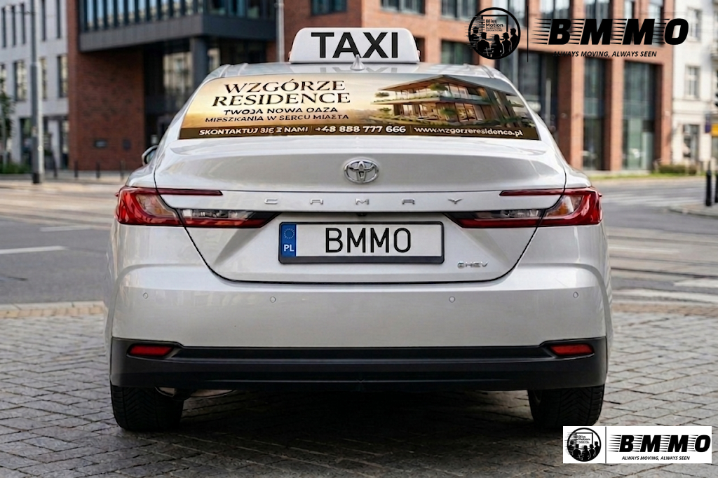 Rear view of a white Toyota Camry taxi with a personalized license plate 'BMNO', parked on a city street. There is an advertisement on the back window promoting Wzgórza Residence, a residential development in Poland.