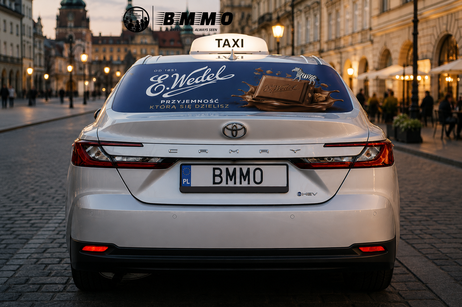 Rear view of a white Toyota Camry electric vehicle with a taxi sign on top, parked on a cobblestone street in an urban area during dusk, with buildings and streetlights in the background.