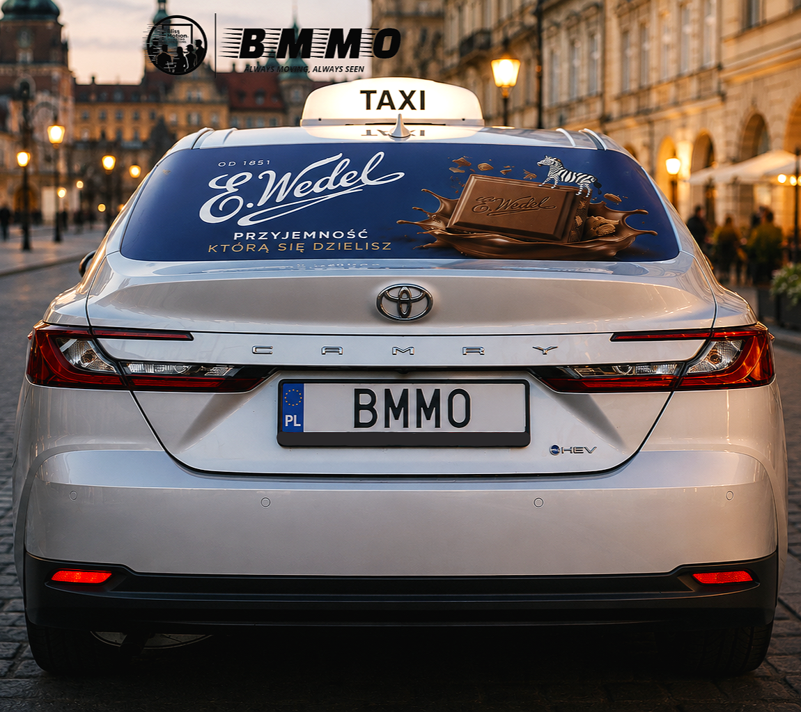 Rear view of a white Toyota Camry car with a taxi sign on top, parked on a city street at dusk. The back window has an advertisement for E. Wedel chocolates with a chocolate bar and a zebra, written in Polish. The car has a Polish license plate reading 'BMMO'.