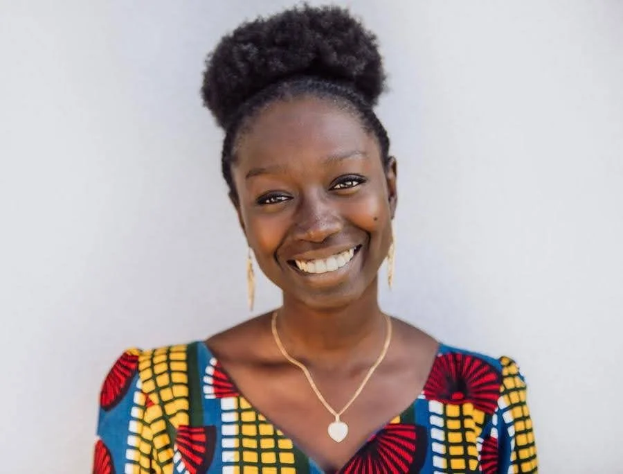 Smiling woman with natural hair styled in a puff, wearing a colorful patterned dress, gold jewelry, and earrings, posing against a plain light-colored background.