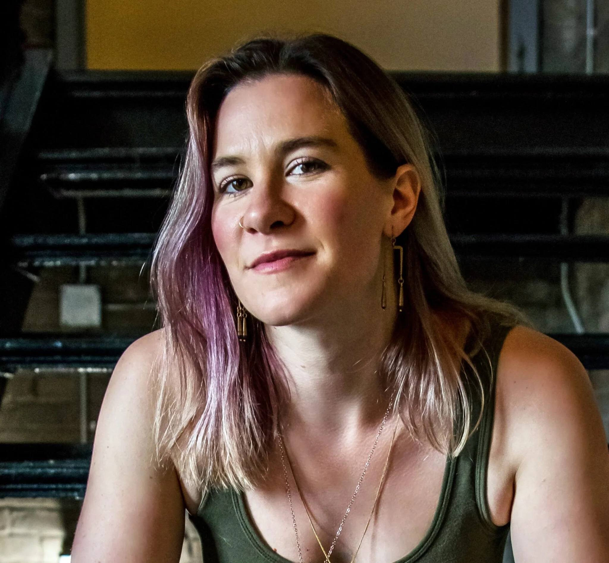 A woman with shoulder-length hair dyed with purple highlights is sitting indoors. She has a nose ring and is wearing gold earrings and layered necklaces. She has a neutral expression and is wearing a sleeveless, dark green top.