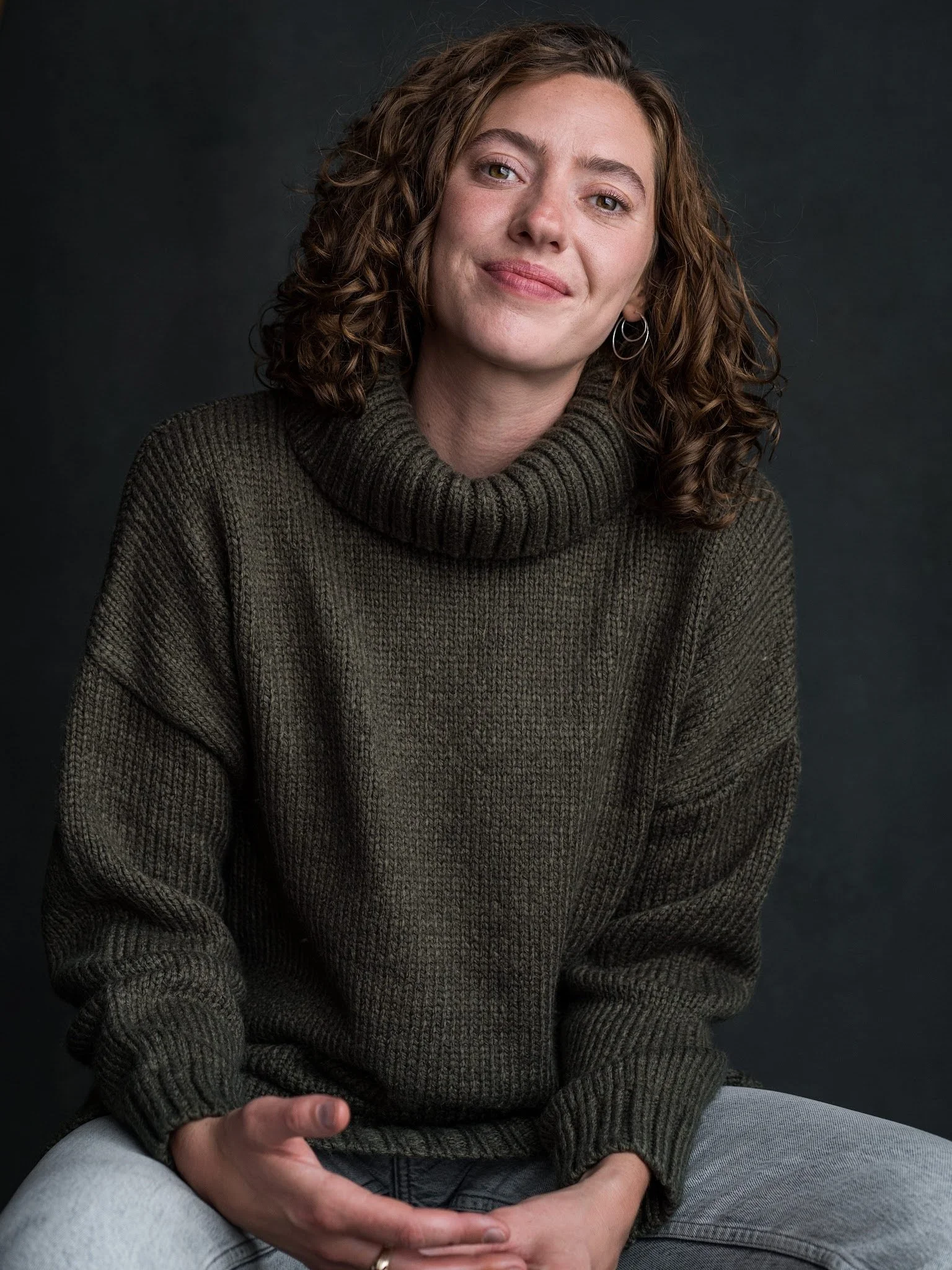 A woman with curly brown hair wearing a dark green turtleneck sweater, sitting against a dark background, smiling softly at the camera.
