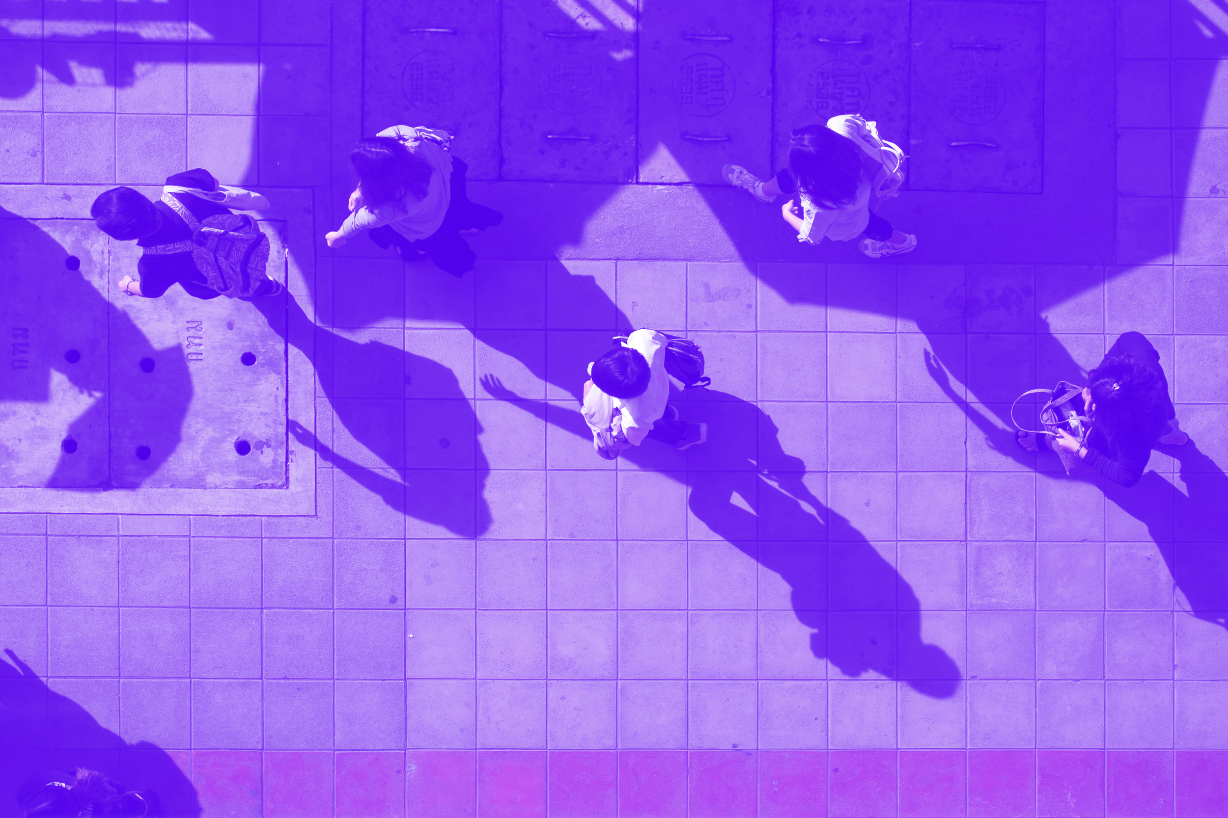 Bird's eye view of seven people walking on a tiled sidewalk, with their long shadows cast across the ground.