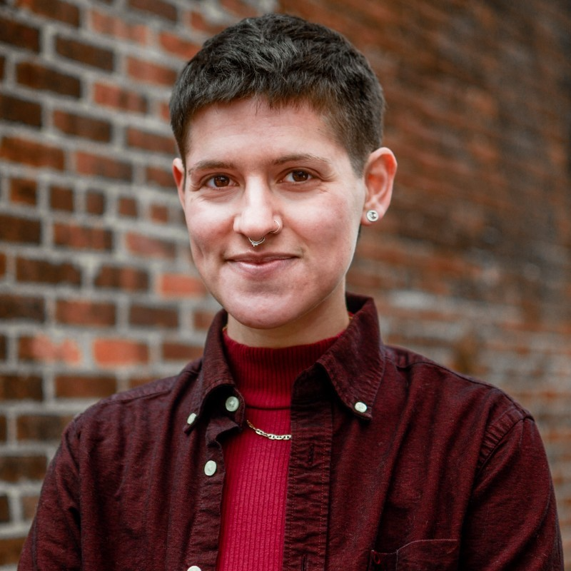 Portrait of a person with short dark hair, multiple piercings including a nose ring and earrings, wearing a red turtleneck and burgundy shirt, standing in front of a brick wall.