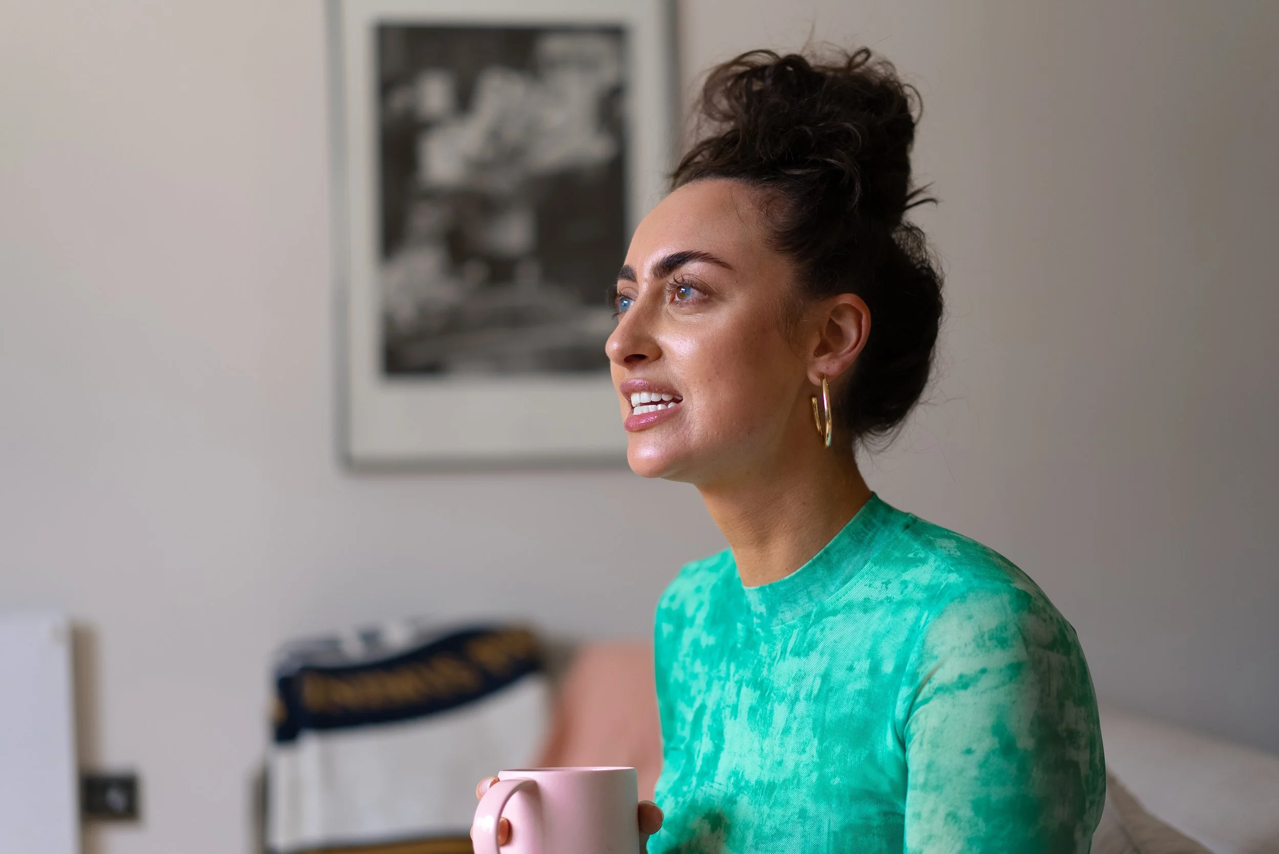 A woman with curly brown hair tied up, wearing a green tie-dye shirt and gold hoop earrings, sitting indoors holding a pink mug, looking to the side.
