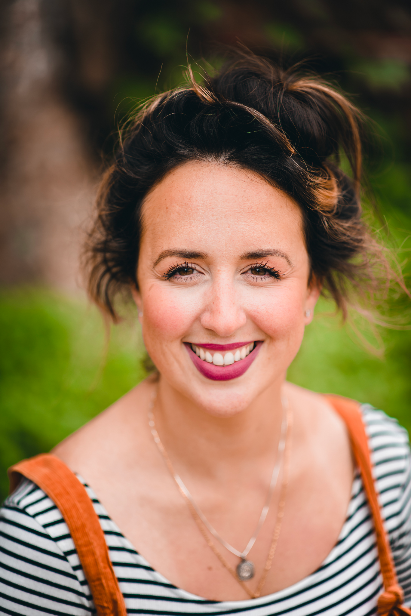 A young woman smiling outdoors, with short dark hair, wearing a striped top, orange suspenders, and layered necklaces, standing against a background of green foliage.