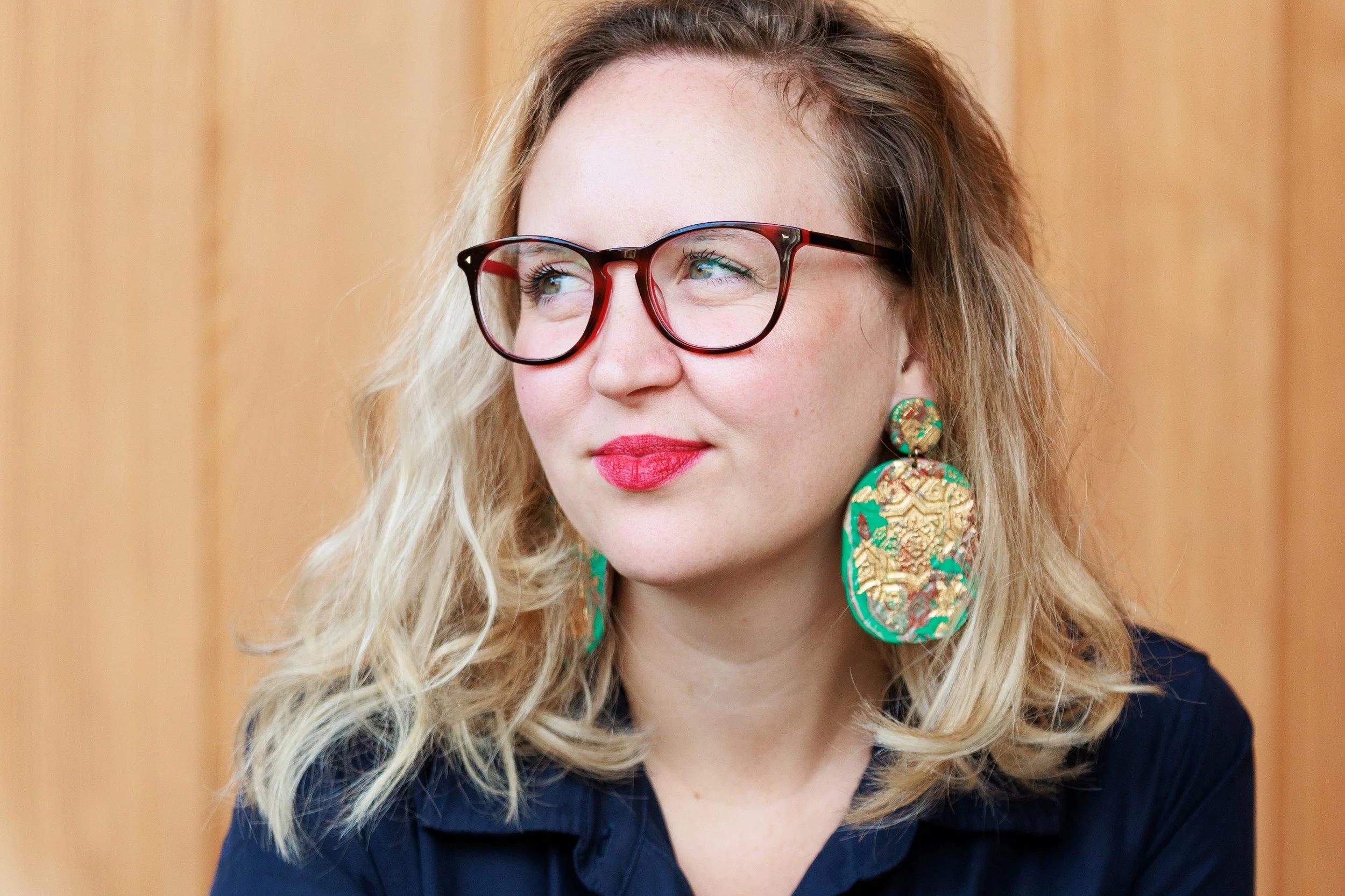 Close-up of a woman with wavy blonde hair, wearing large green and gold earrings, red glasses, red lipstick, and a dark top, against a wooden background.