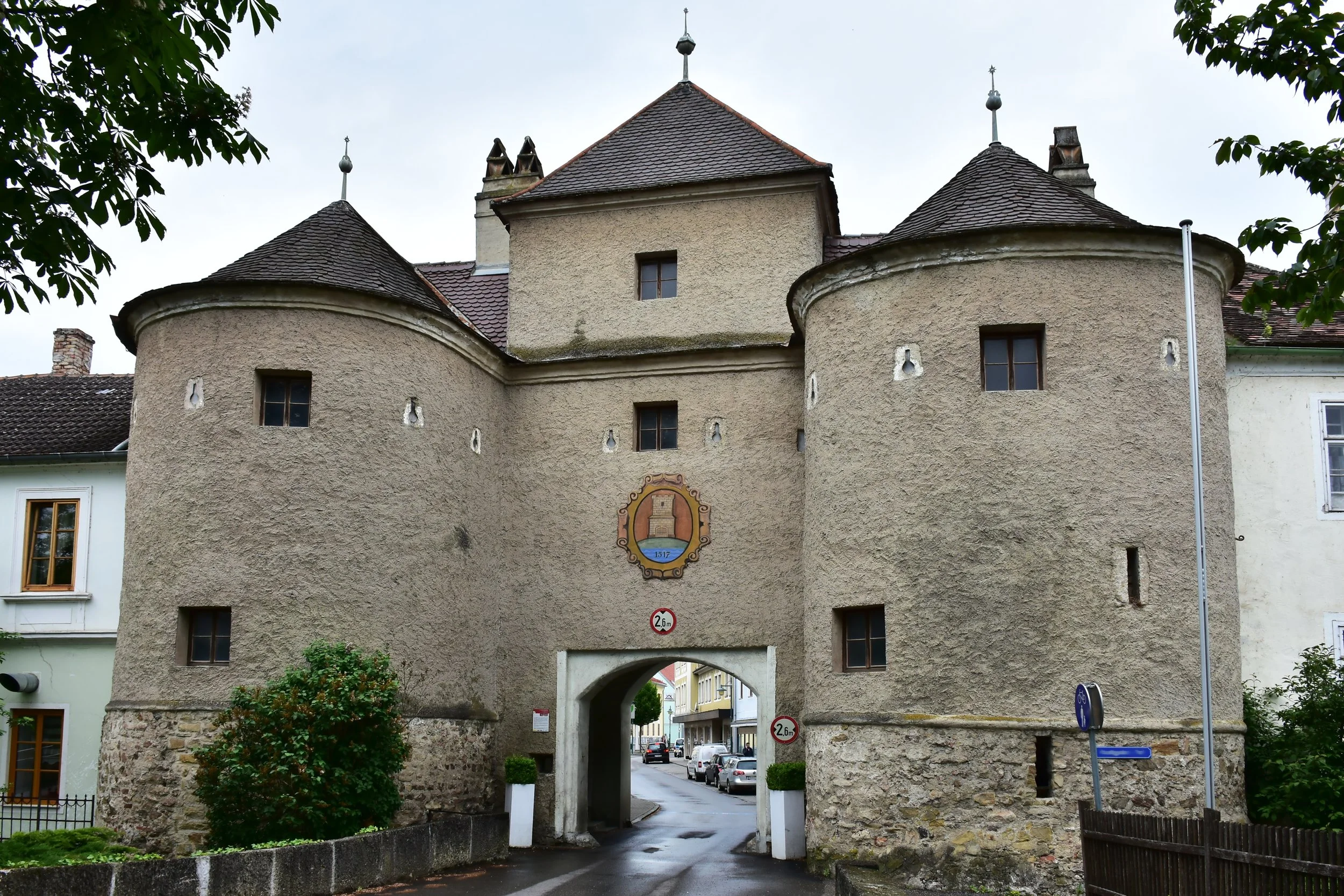 Old stone and stucco city gate with an arched passageway, small windows, decorative crest with a tower symbol, and three peaked rooftops with finials.
