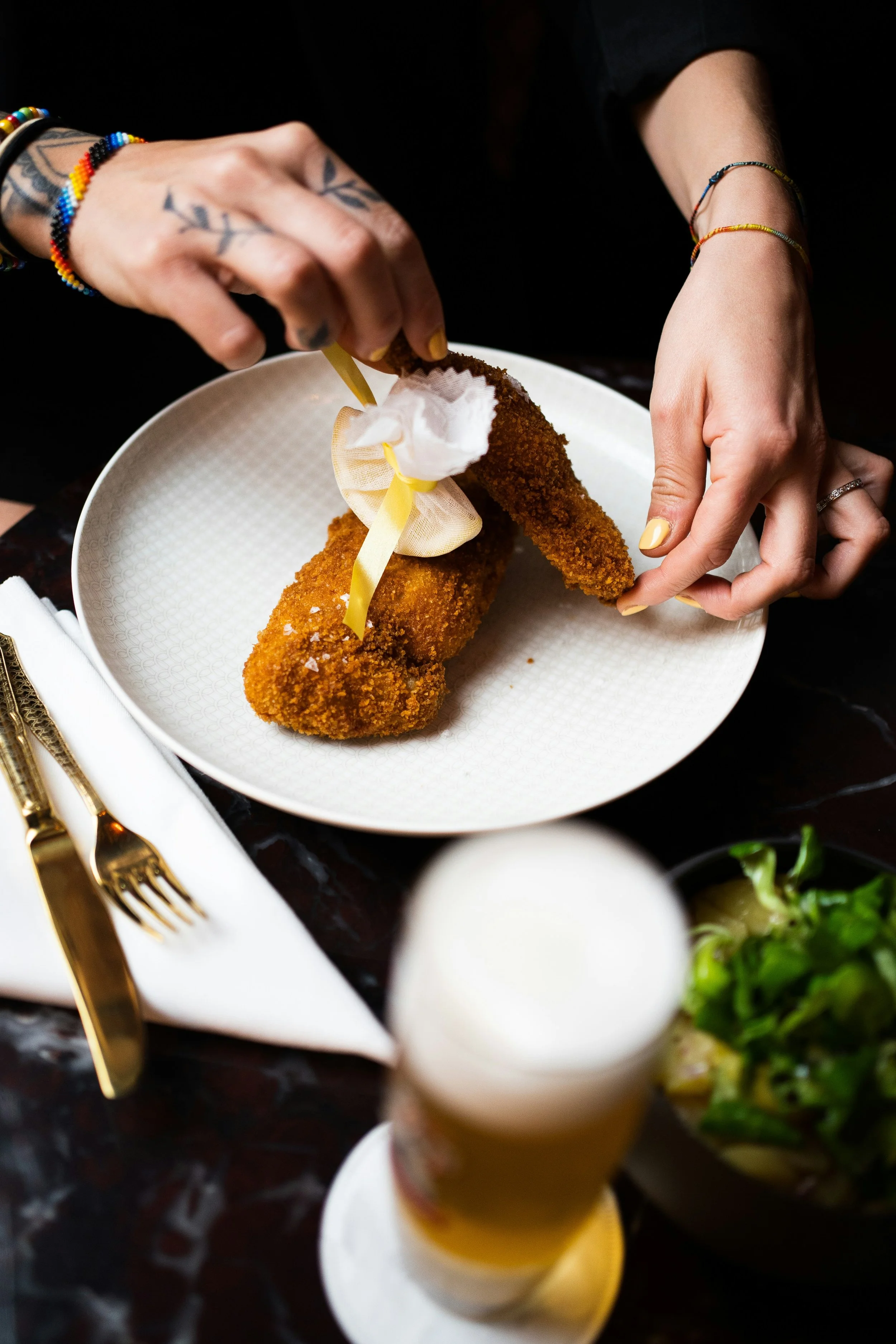 A person preparing a breaded fried fish fillet with a decorative ribbon around it on a white plate, surrounded by small bowls and utensils on a dark tabletop.