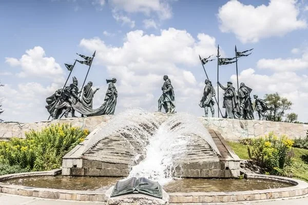 A statue of soldiers with flags standing on a hill, with water cascading down a stepped platform and forming a small waterfall, in front of a cloudy sky.