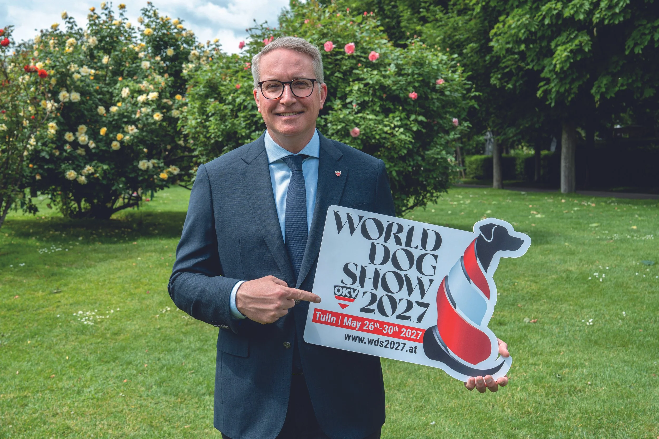 A man in a suit and glasses standing outdoors in front of bushes with flowers holding a sign promoting the World Dog Show 2027 in Tulln, Austria, scheduled for May 26th-30th, 2027.