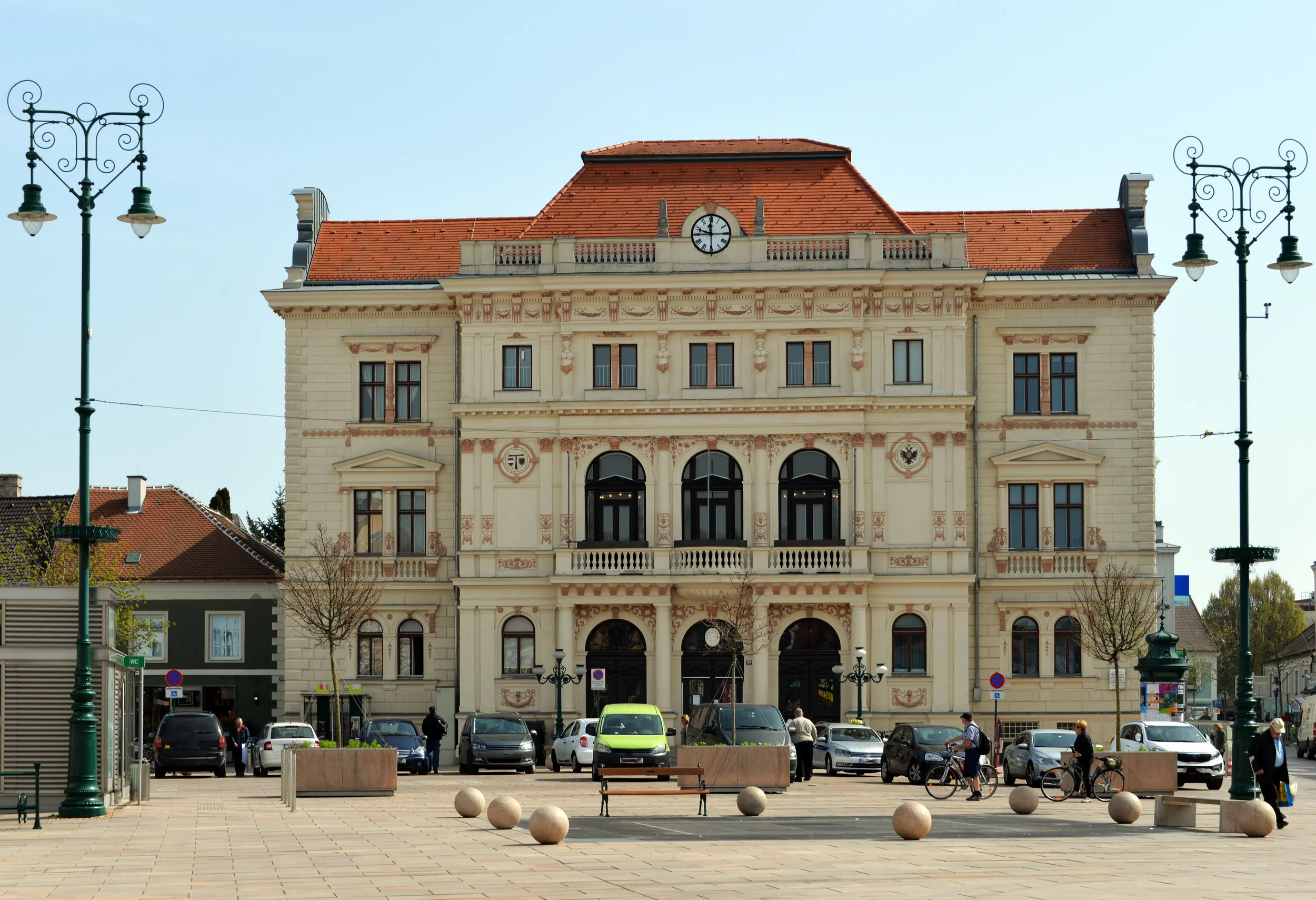Front view of a historic building with a clock on the roof, red tiled roof, and ornate architectural details. There are lampposts on either side and a parking lot with people walking and cycling in the foreground under a clear blue sky.