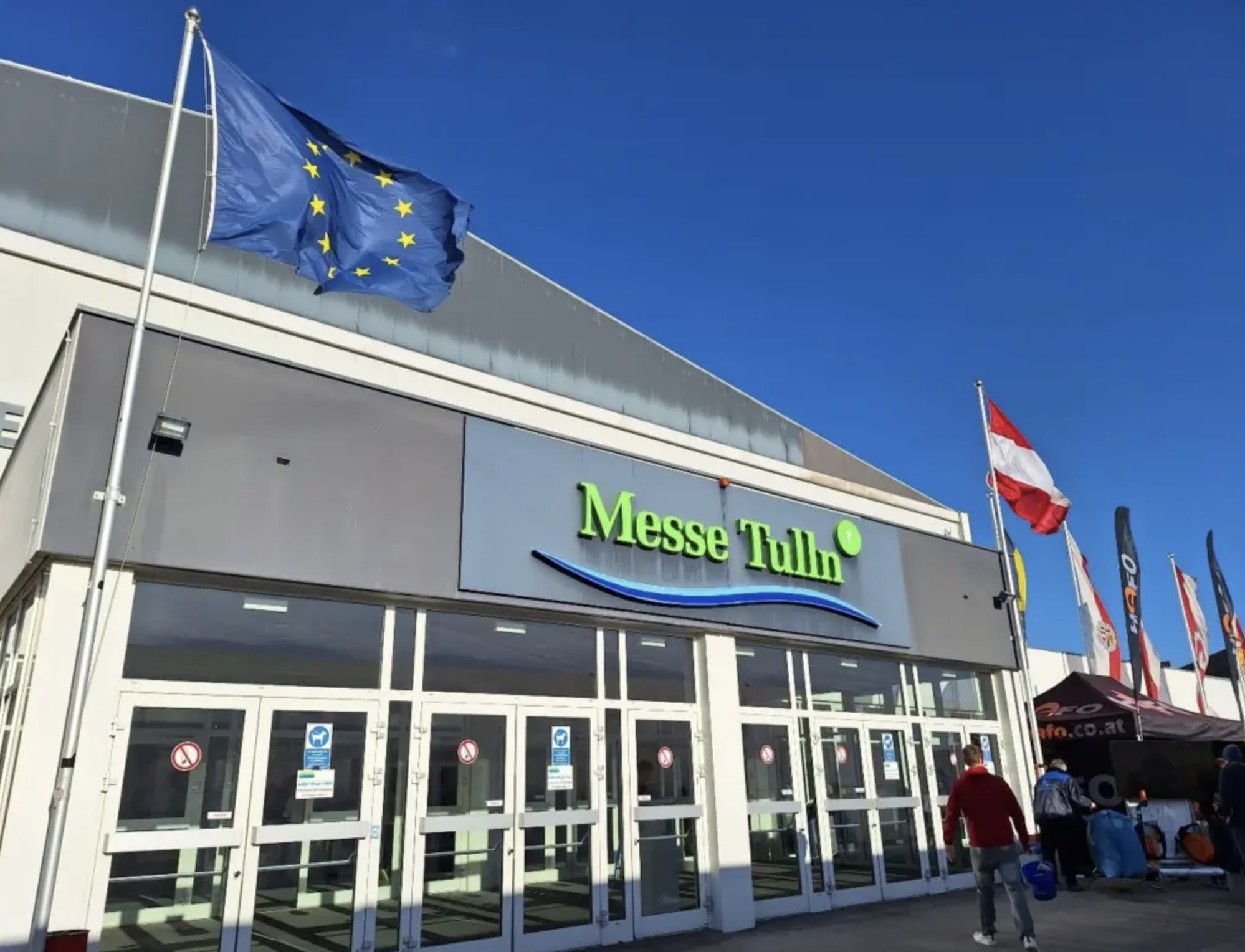 Exterior of a Messe Tulln exhibition center with flags, including the European Union flag and the Austrian flag, against a bright blue sky. A few people are walking near the entrance.