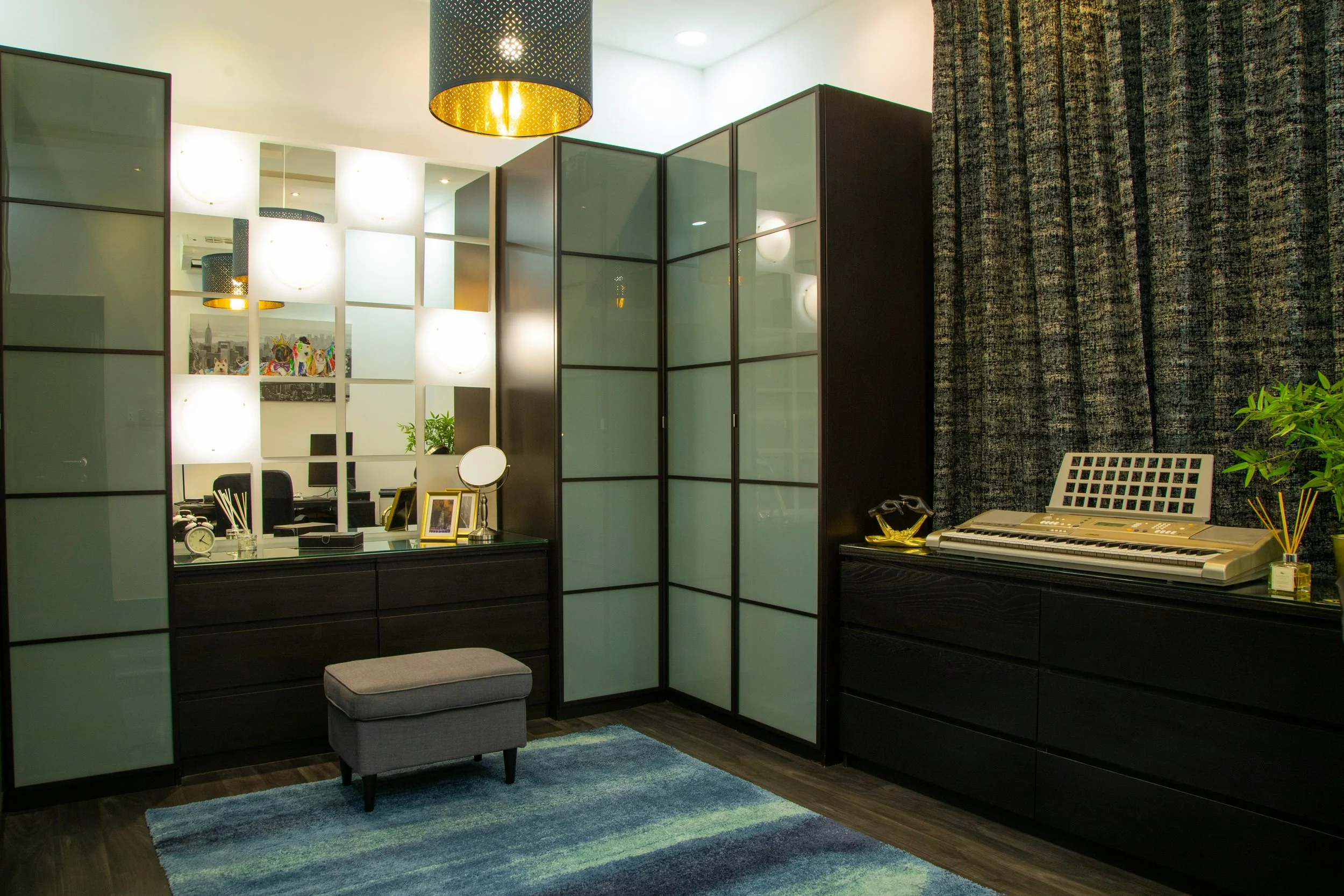 Interior of a modern room with glass-paneled room dividers, a desk with decorations including framed pictures and a mirror, a gray ottoman, a colorful rug, and a black digital piano, against a textured dark curtain background.