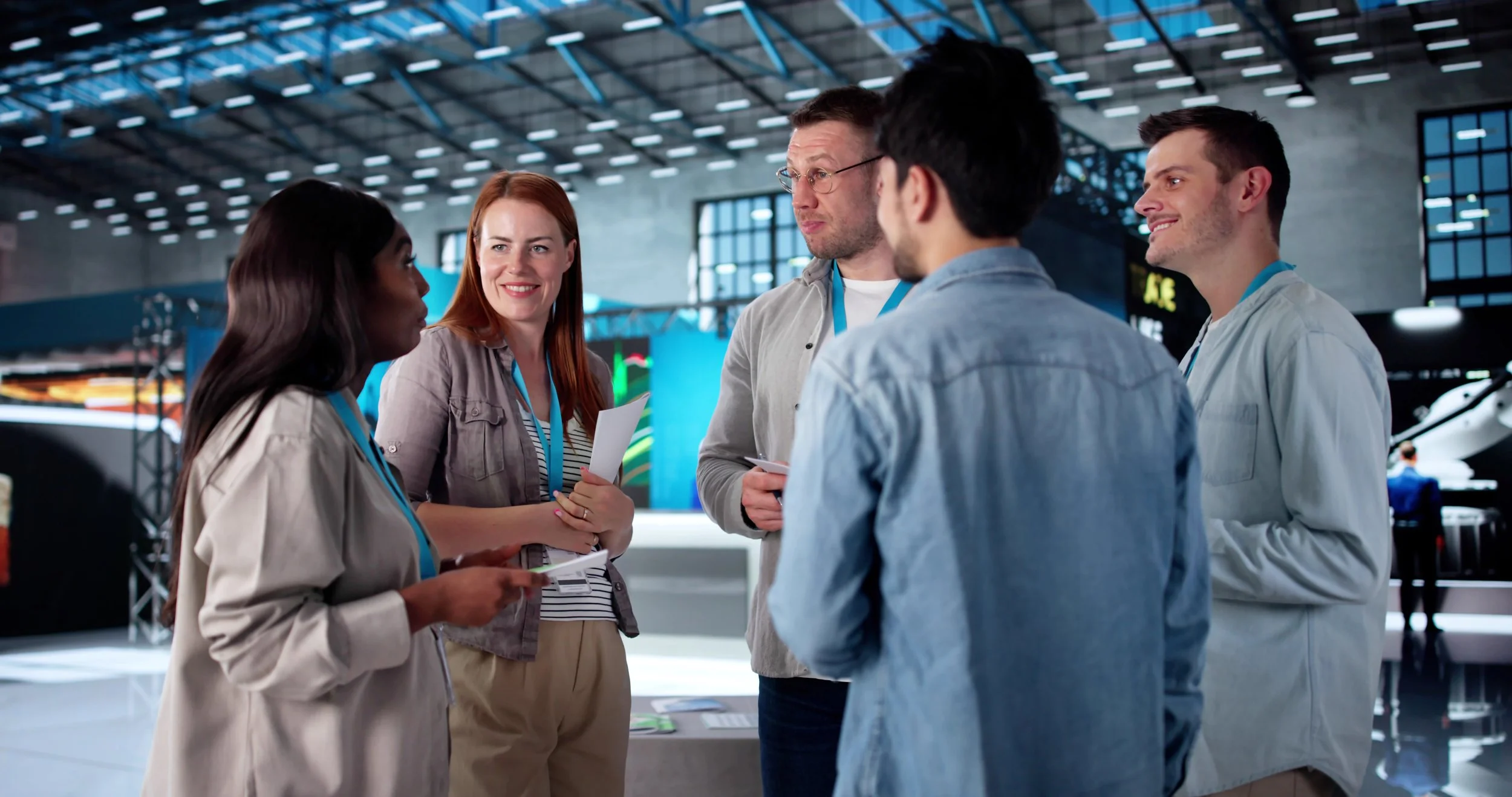 Group of five people having a discussion at an event or conference in a large indoor venue with high ceilings and large windows.