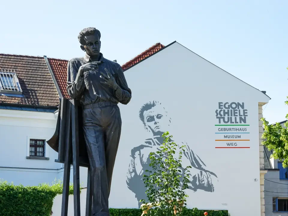 A bronze statue of a woman standing outdoors in front of a white building with a mural of her face. The building's wall has text that reads 'Egon Schiele Tulln' and lists a birth house, museum, and WEG.