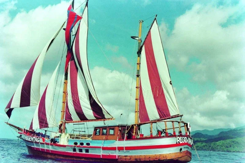 A large wooden sailing ship with pink, purple, and white sails on the ocean, under a partly cloudy sky.