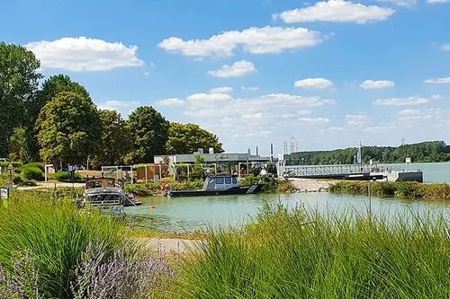 A boat docked at a marina near a river or lake, with trees, a bridge, and a partly cloudy sky in the background.