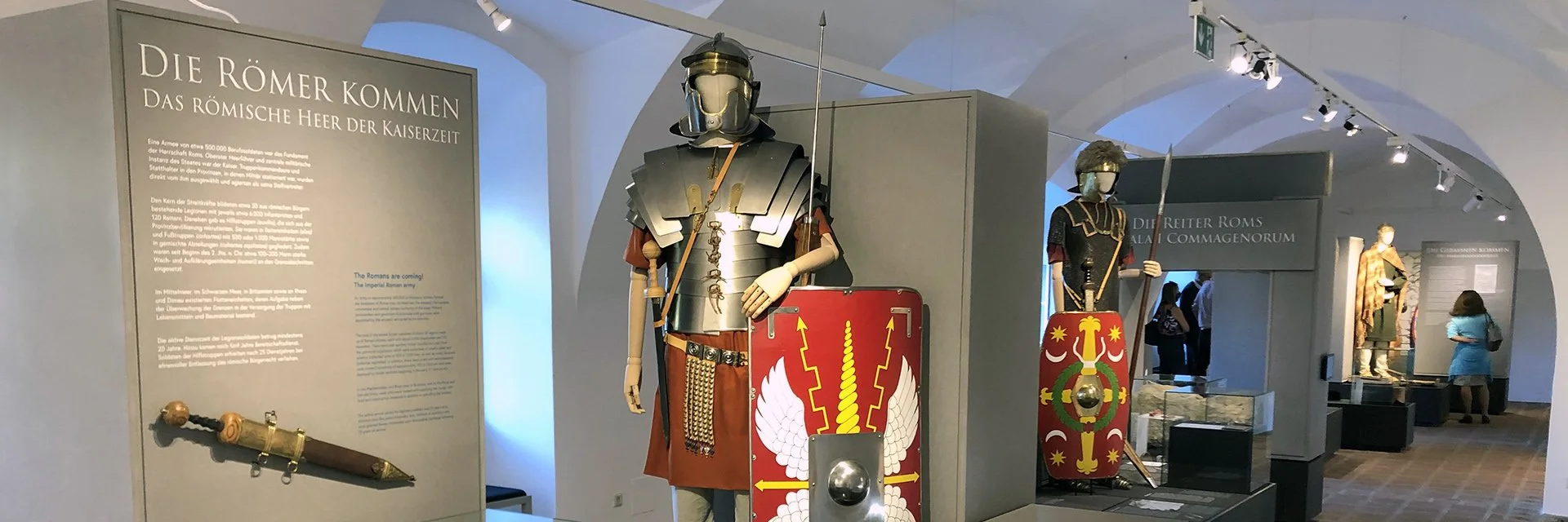 Museum exhibit featuring Roman legionary armor and shields, with informational panels and display cases in the background.