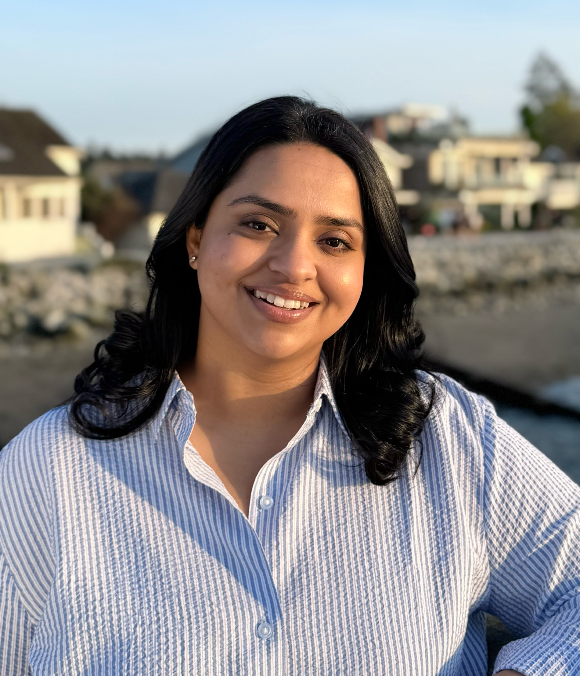 Diksha Tyagi - Counsellor - A woman with black hair and a striped shirt smiling outdoors near water with houses in the background.