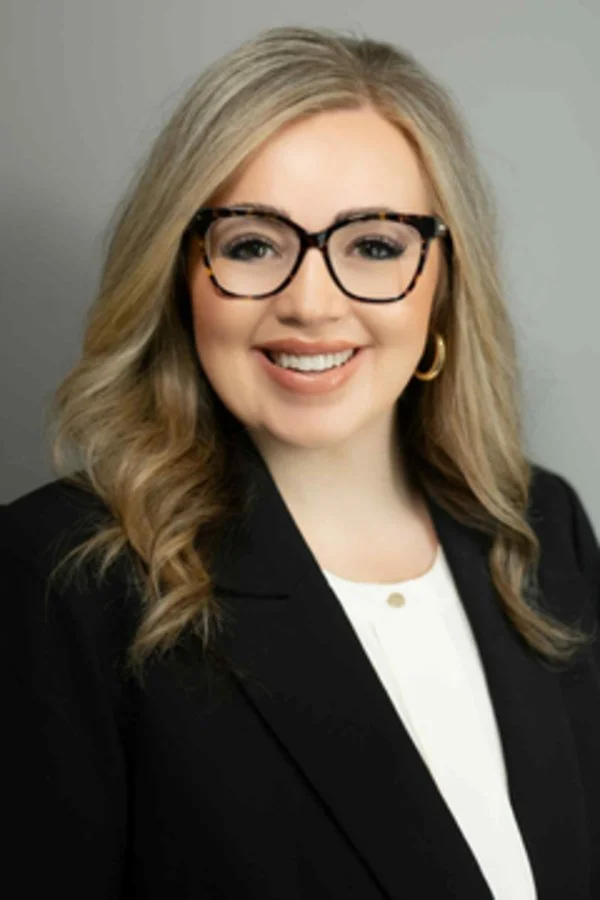 A woman with blonde hair, glasses, earrings, wearing a black blazer over a white top, smiling at the camera.