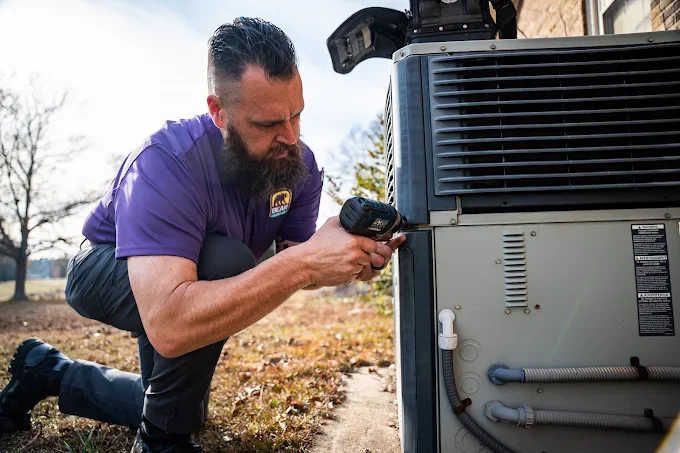 A technician wearing a purple shirt kneels outdoors, using a diagnostic tool on an air conditioning unit.