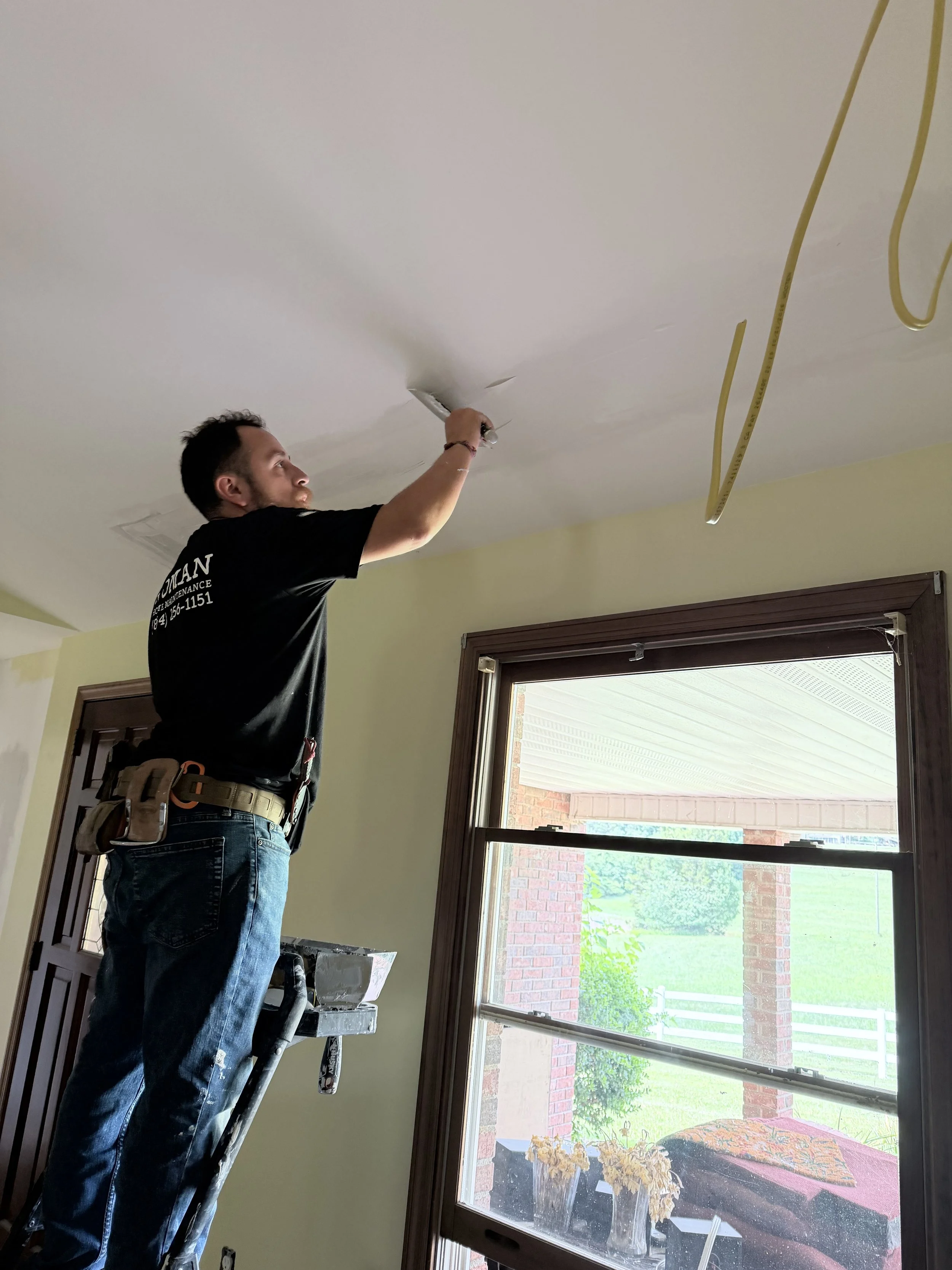 A man standing on a ladder repairing a ceiling in a room, with a large window revealing a patio and garden outside.