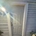 Exterior door with white framing and part of a blue house siding visible, sunlight shining on the door area