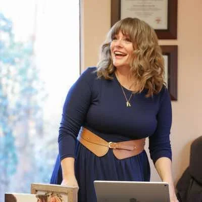 A woman with shoulder-length curly hair, wearing a dark blue dress with a beige belt, smiling in an office setting.
