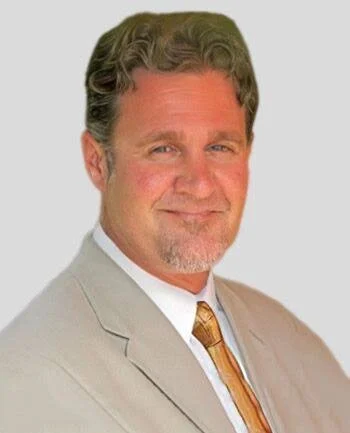 A middle-aged man with curly hair and a beard, wearing a light beige suit, white shirt, and patterned tie, smiling confidently.