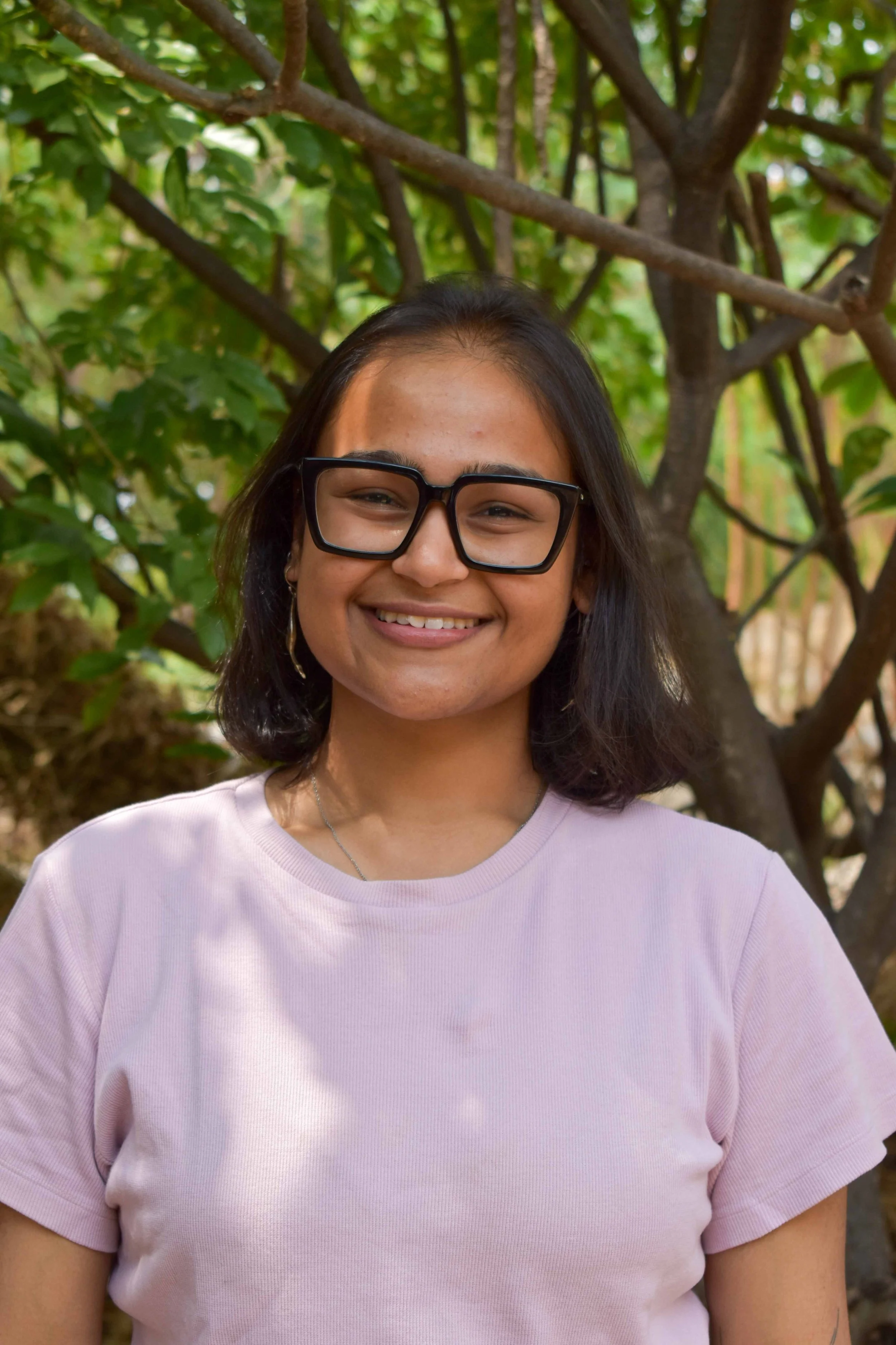 A young woman with shoulder-length dark hair, wearing glasses and a light pink t-shirt, smiling outdoors in front of green foliage and a tree.