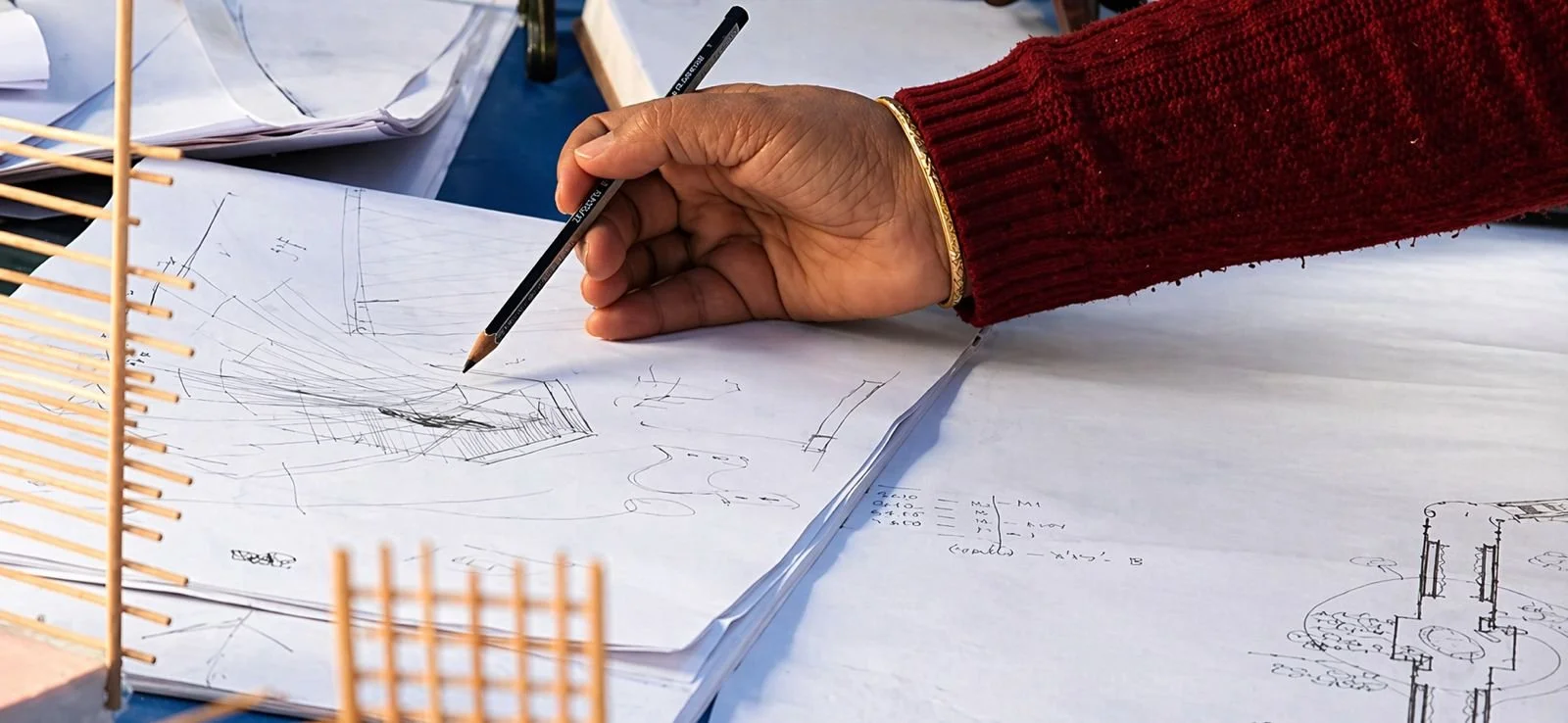 A person in a red sweater is drawing architectural sketches on paper with a pencil, with a model structure and additional papers visible on the table.