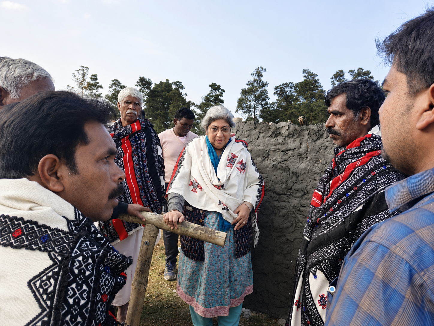 A woman in traditional attire speaks with several men in traditional patterned shawls outdoors, with trees and a stone wall in the background.