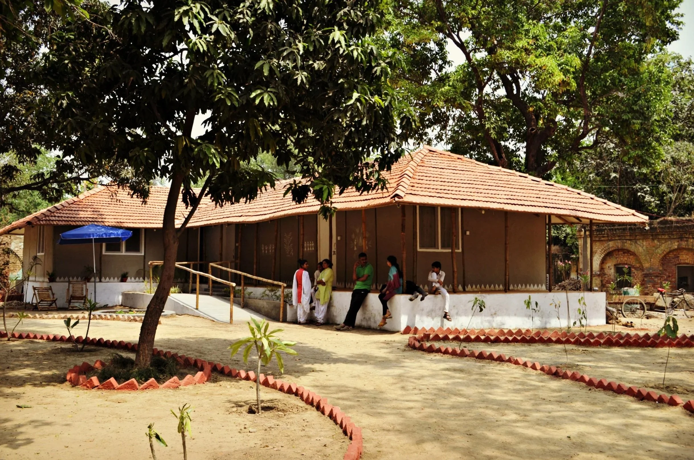 A group of people standing on the porch of a building with a red-tiled roof, surrounded by trees and a courtyard with a pathway and small plants.