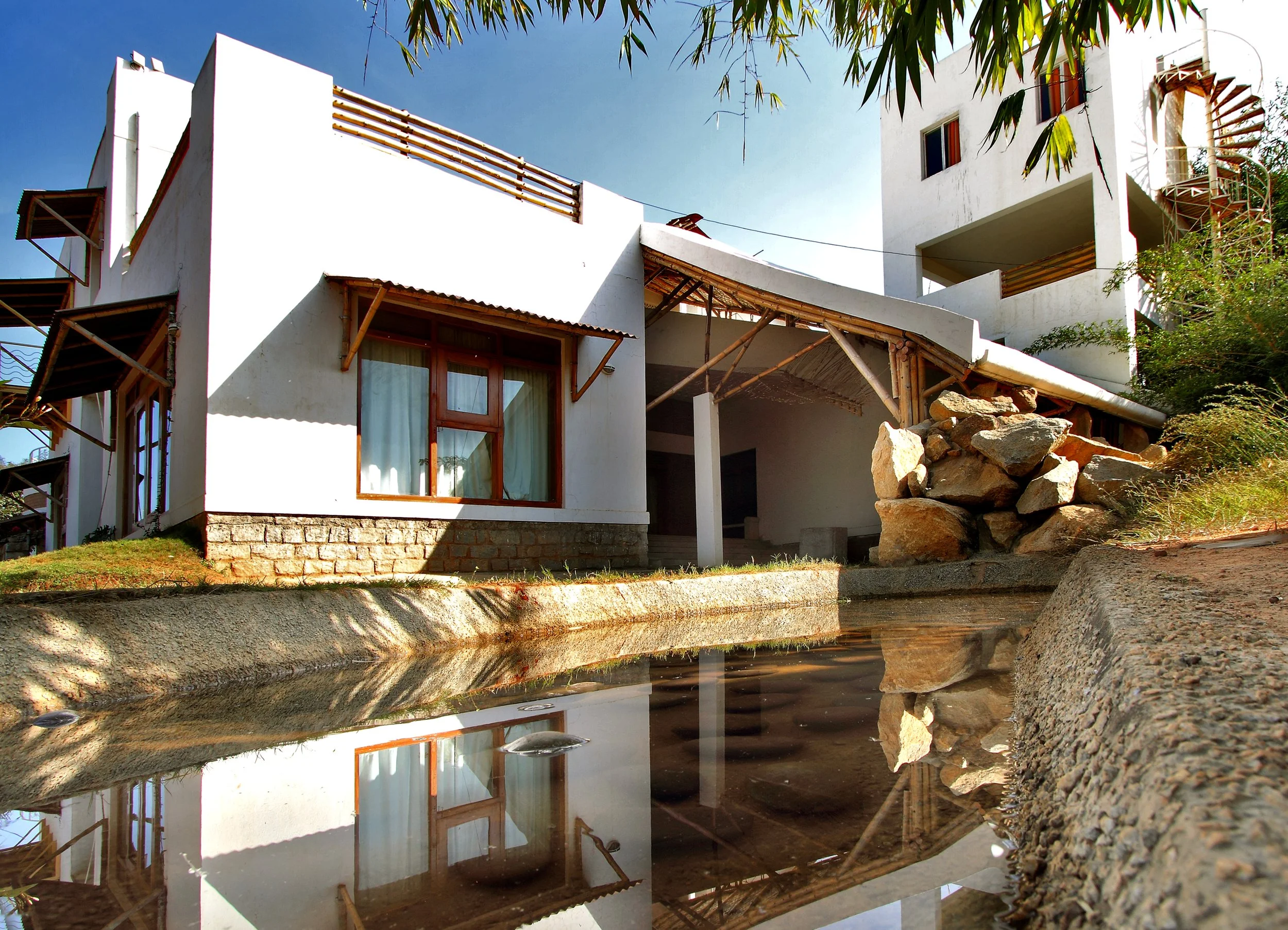 Modern white multi-story house with wooden accents and large windows, situated beside a small body of water reflecting the building, under a clear blue sky with some green foliage.