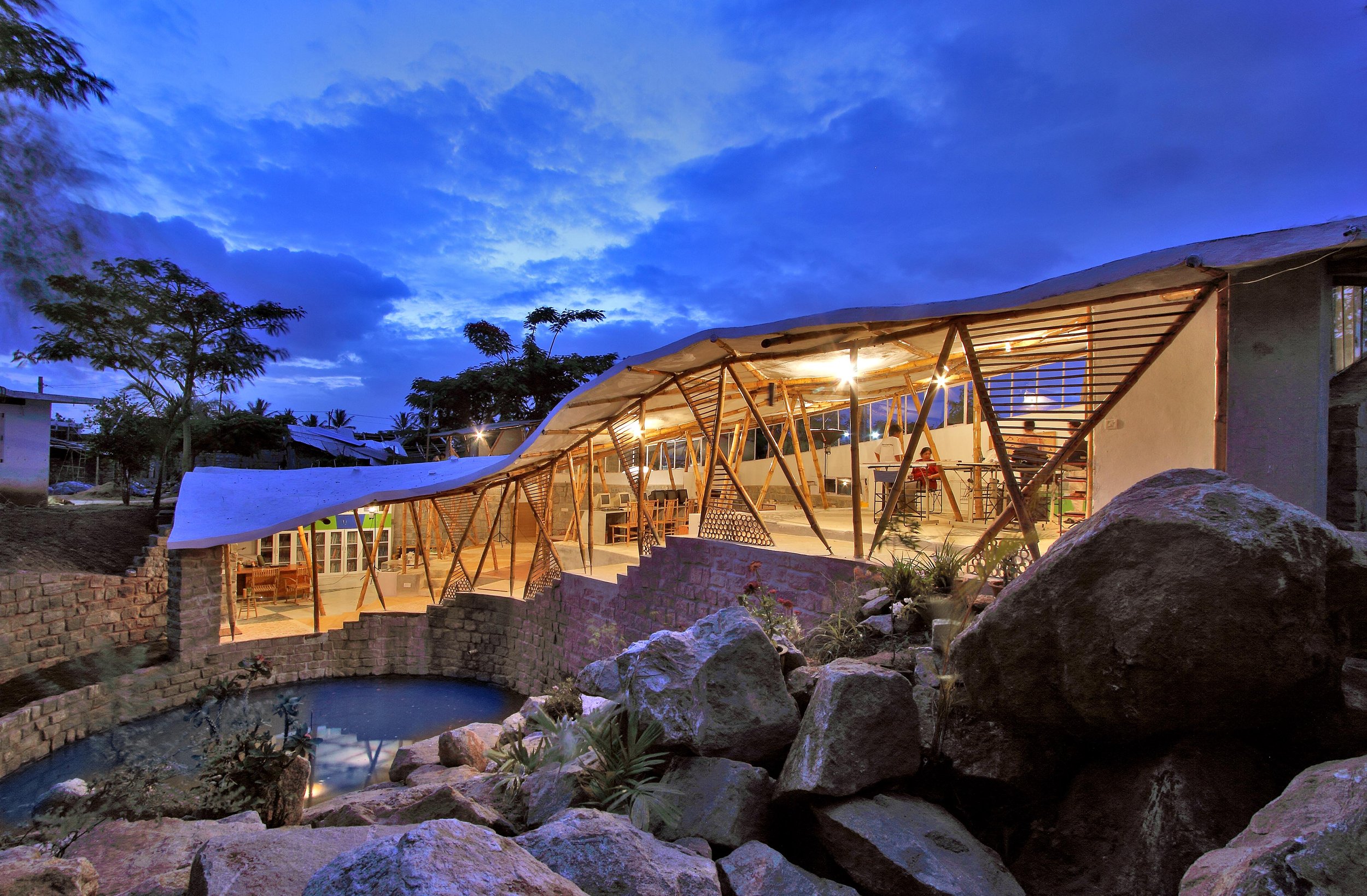 An outdoor restaurant or café with a modern design, featuring a curved bamboo and thatch roof, illuminated by string lights. There are seating areas with tables and chairs, surrounded by rocks and a small pond, against a backdrop of trees and a blue evening sky with clouds.