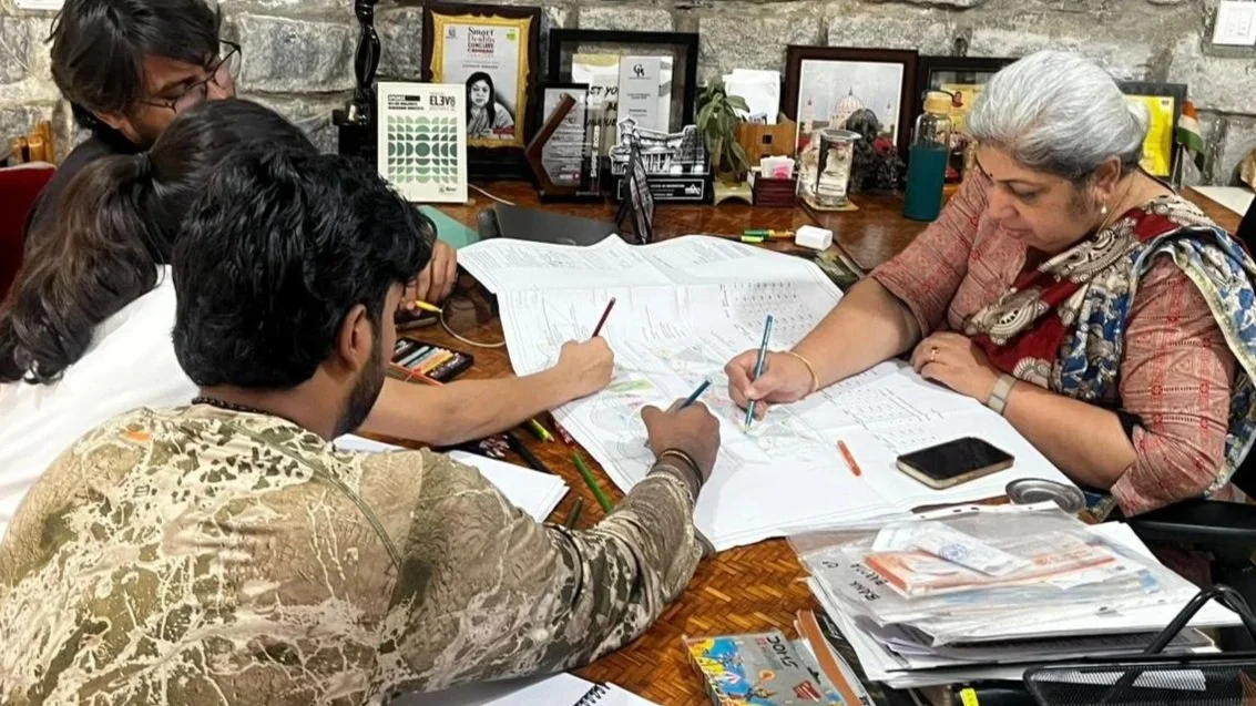 Four people sitting around a table reviewing documents, with writing utensils, a phone, and various office supplies on the table, against a stone wall background.