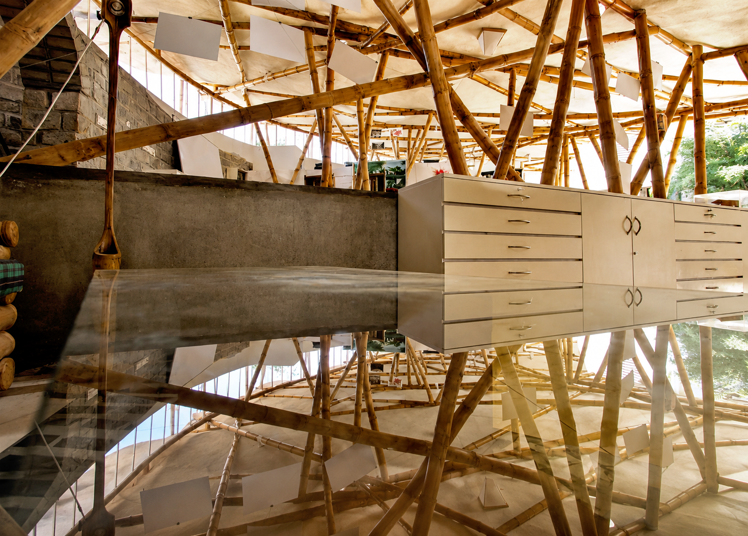 Interior view of a building with a bamboo structure, a concrete wall, and a beige cabinet with drawers, reflected on a shiny surface.