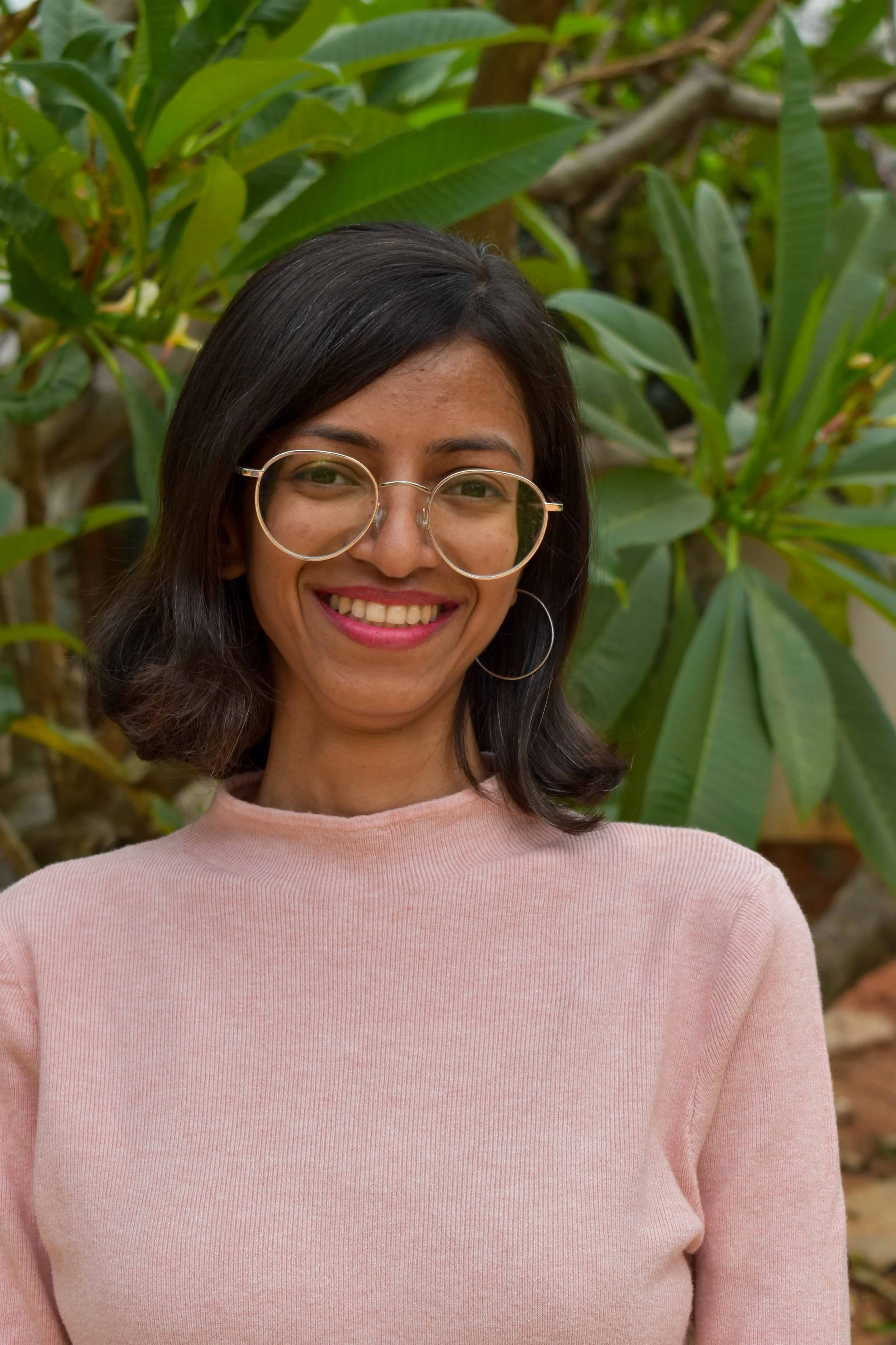 A woman with dark hair and glasses smiling outdoors with green leafy plants in the background.