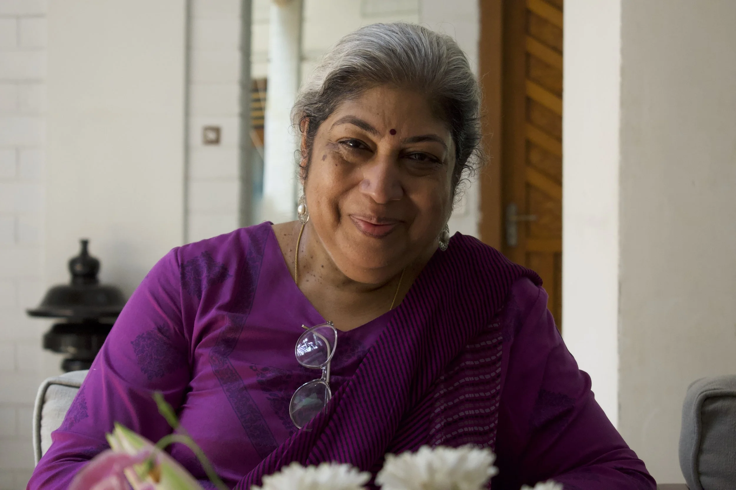 A woman with gray hair, wearing a purple traditional Indian saree, smiling and sitting indoors near a flower arrangement.