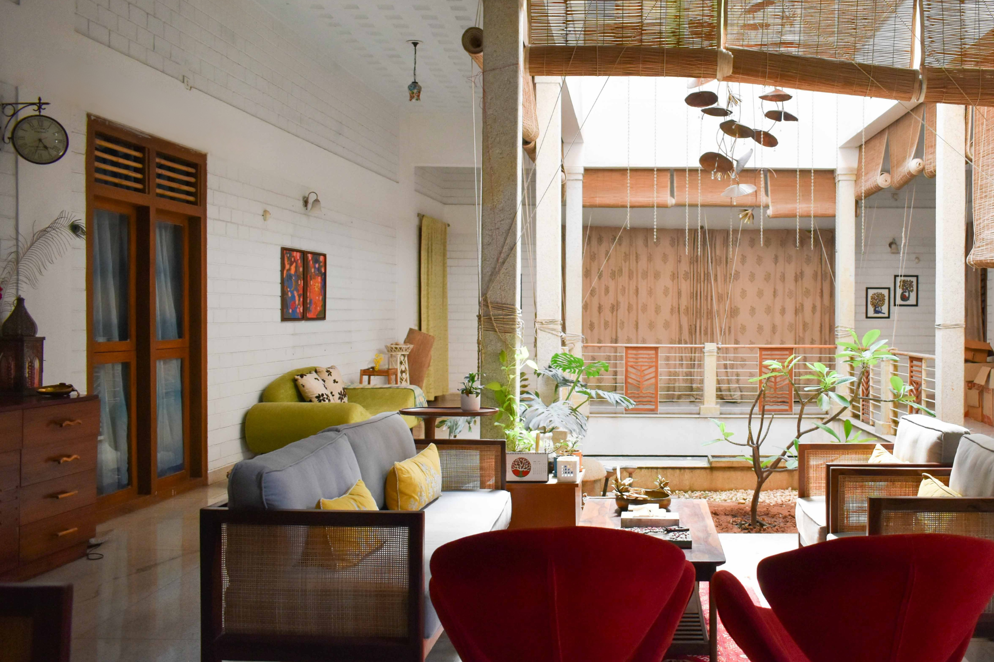 Interior of a cozy living room with white brick walls, a green sofa, red velvet chairs, and various potted plants. Large windows and a balcony with bamboo blinds let in natural light. Decor includes framed artwork and a wall clock.