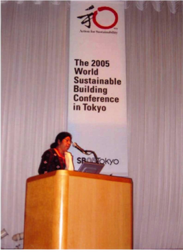 A woman speaking at a podium during the 2005 World Sustainable Building Conference in Tokyo, Japan, with a large vertical banner behind her.