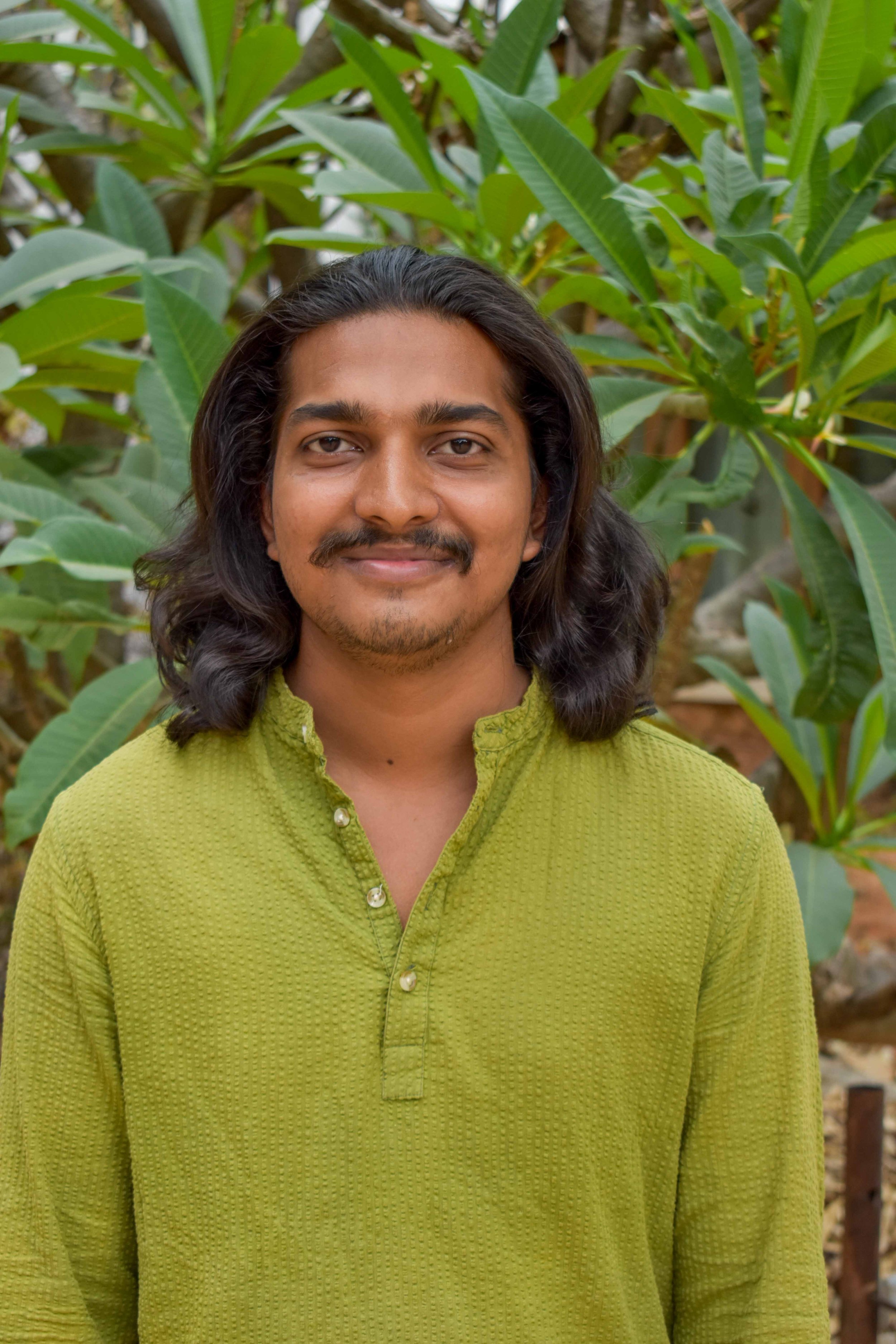 A man with long, dark hair and a slight beard, smiling at the camera, wearing a green kurta, stands in front of a background of large green leaves and plants.
