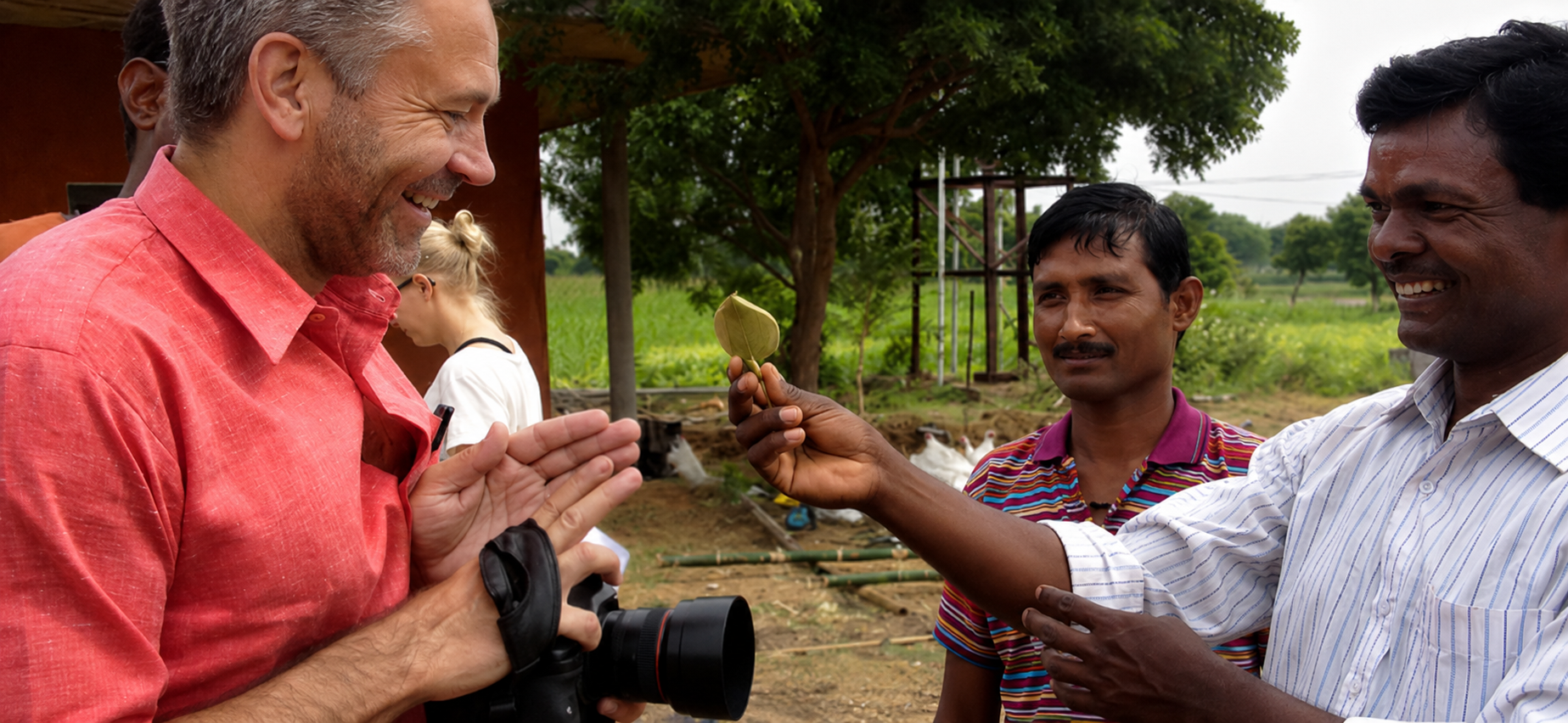 A group of four people outdoors, smiling and engaging in a cultural exchange. A man in a red shirt is facing left, holding a camera and smiling. A man in a striped shirt is holding a leaf and presenting it, while another man in a white striped shirt is smiling. A woman in the background is looking down. Trees and farmland are visible in the background.