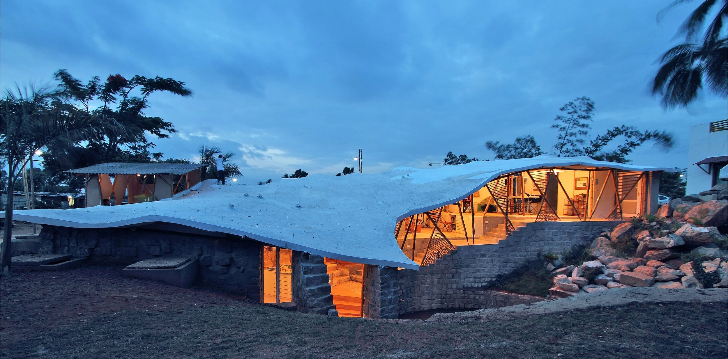 A modern building with a curved, white roof and illuminated interior, set among trees and rocks during dusk or early evening.