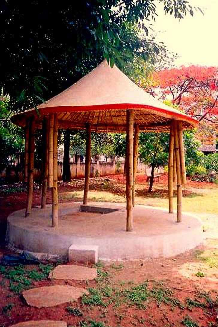 A small gazebo with a conical thatched roof supported by wooden poles, situated on a round concrete platform in a garden surrounded by trees and foliage.