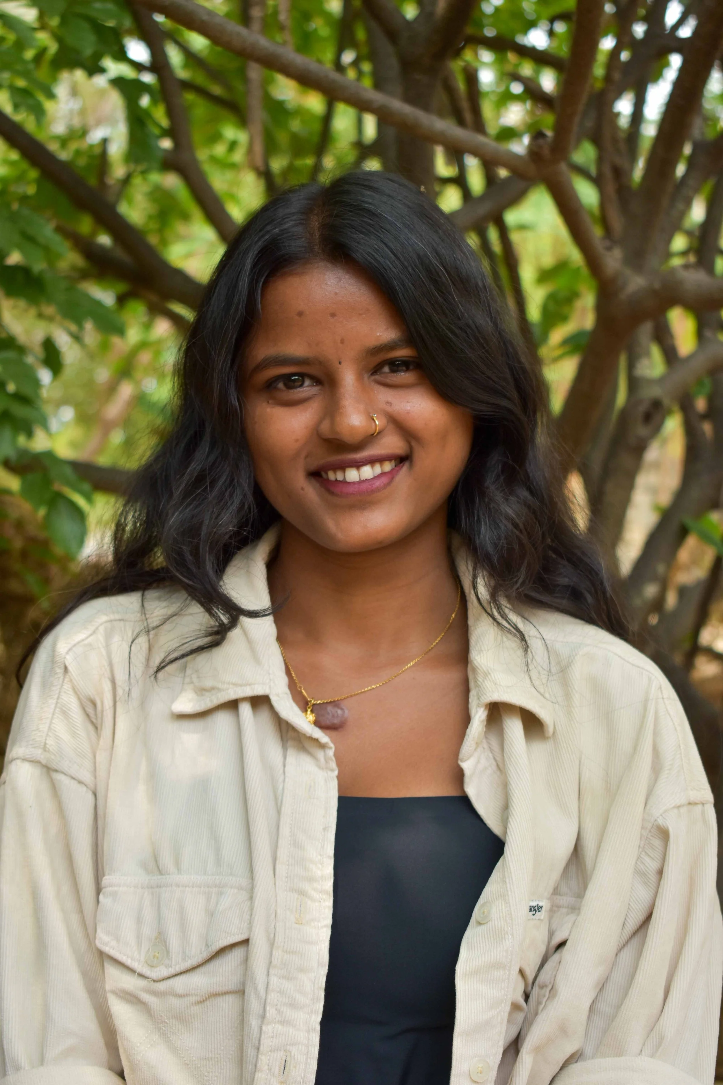 A smiling woman with black wavy hair standing outdoors in front of green foliage and tree branches, wearing a beige jacket over a black top.