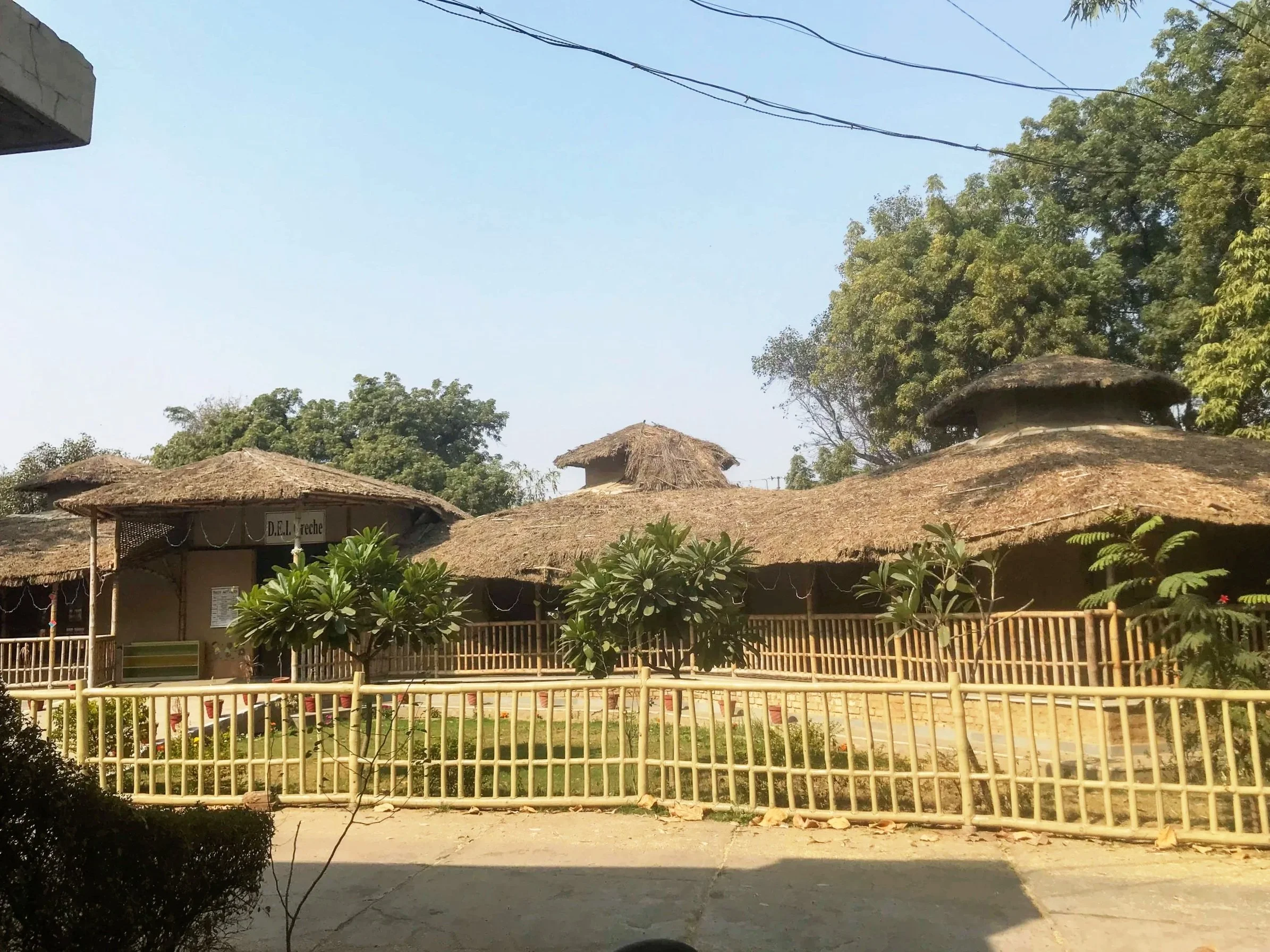 A school yard with traditional round thatch-roof huts, a sign with Spanish text, and a wooden fence in the foreground, surrounded by lush green trees under a clear blue sky.