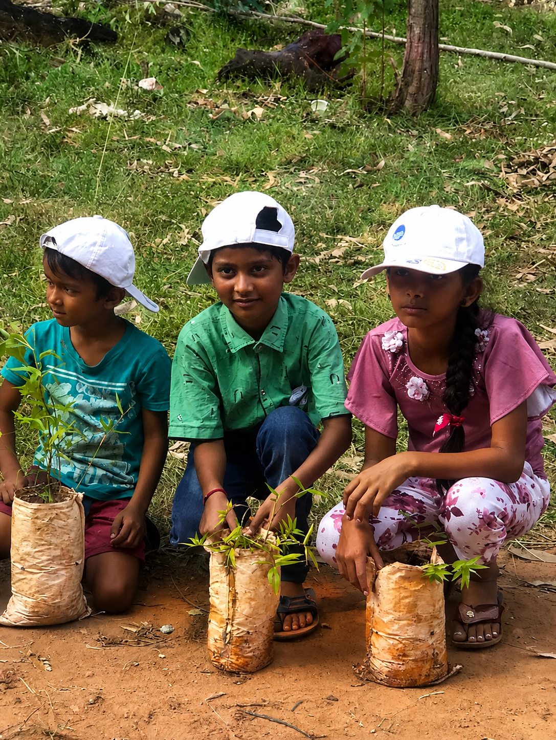 Three children squatting on the ground outdoors, each holding a potted plant, with a forested background and a stray animal in the distance.