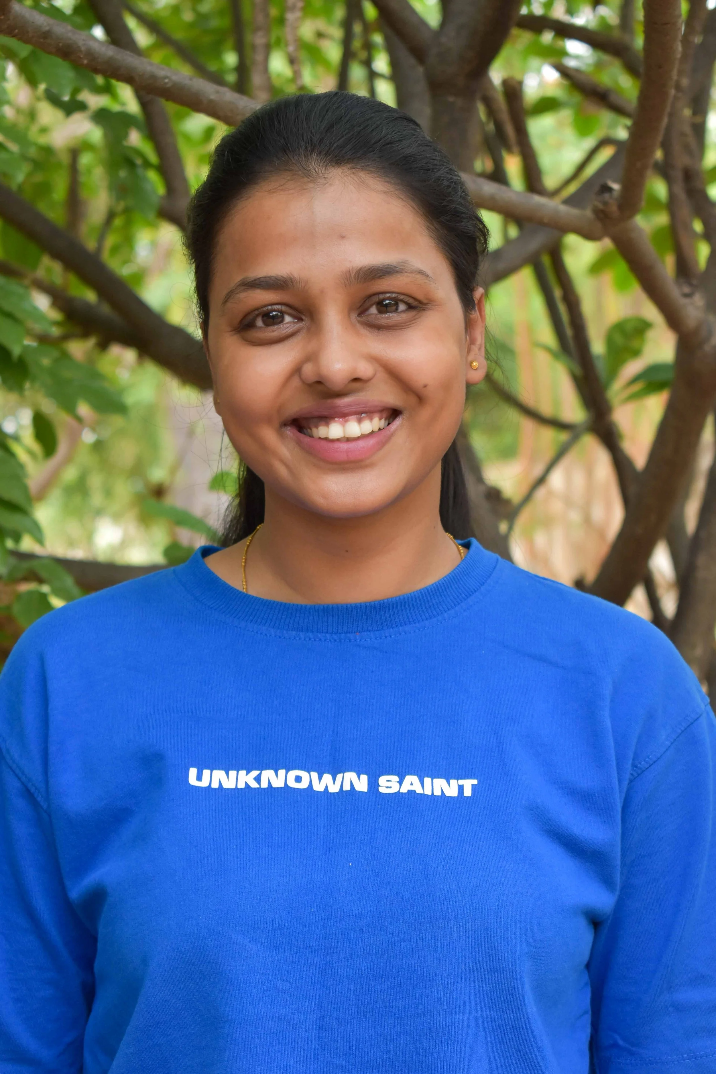 A young woman with dark hair pulled back, smiling, wearing a blue sweatshirt with white text that reads "UNKNOWN SAINT," standing outdoors among green foliage and tree branches.